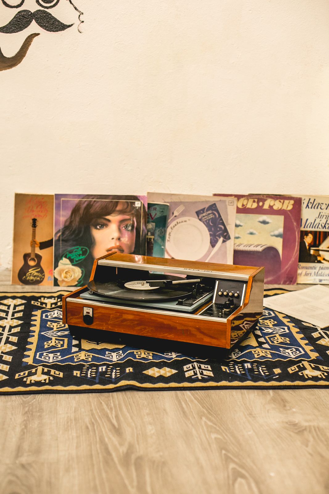 A vintage turntable on a patterned rug, surrounded by vinyl records and a guitar.