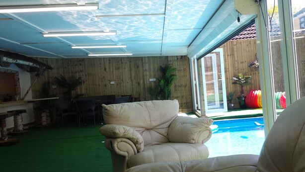 A room with a green carpet, a beige sofa, a dining table with chairs, and a view of a swimming pool through a glass door. - Tudor House (Wisbech)