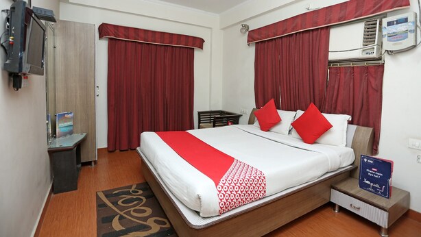 A hotel room with a bed, red curtains, a ceiling fan, an air conditioning unit, a small bedside table, and a digital clock displaying a message. - OYO 12682 Hotel Relax (Gwalior)