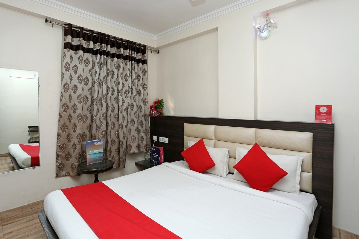 A hotel room with a bed, a nightstand, a mirror, a window with curtains, and a wall-mounted light fixture.