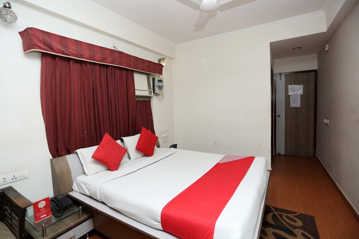 A hotel room with a bed, red curtains, a bedside table with a phone, and a wall-mounted air conditioner.
