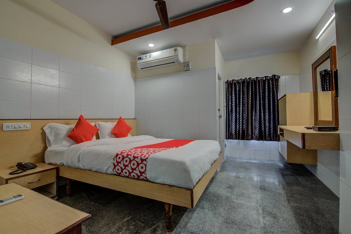 A hotel room with a bed, a bedside table with a phone, an air conditioning unit, a bathroom with a shower, and a mirror.