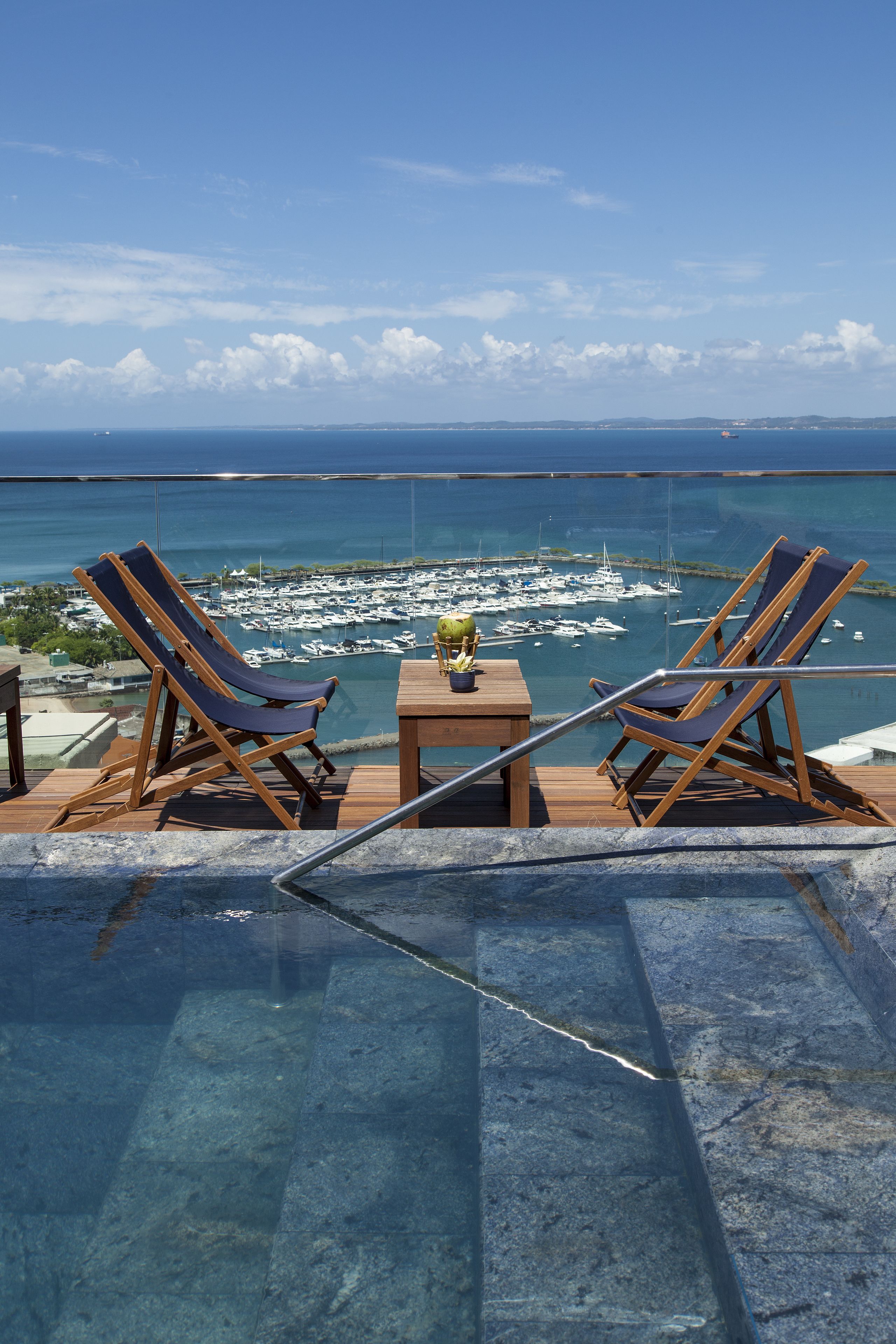 Área à beira da piscina com duas espreguiçadeiras e uma pequena mesa, com vista para um porto cheio de barcos e um céu azul límpido.