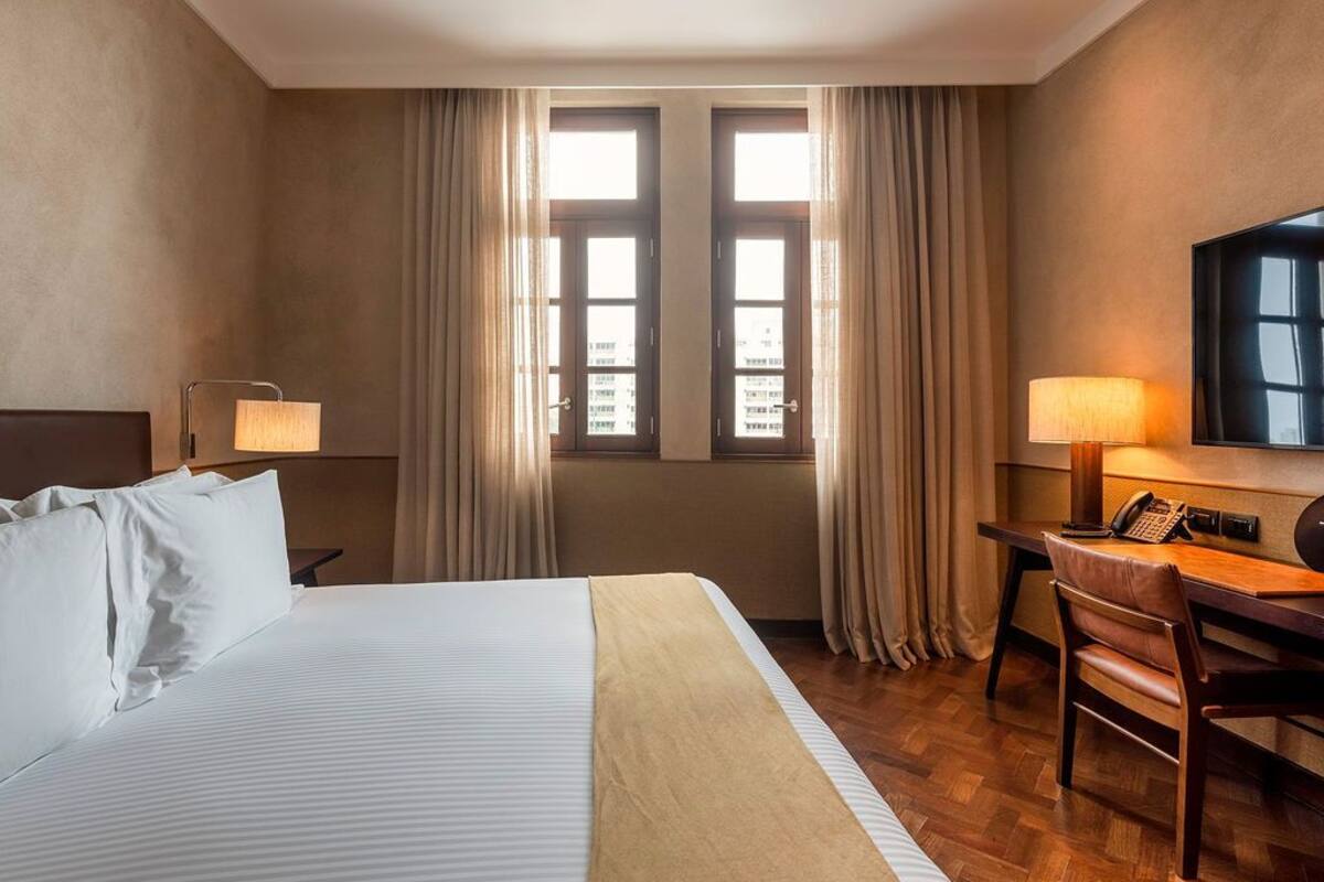 Ein Hotelzimmer mit einem Bett, einem Schreibtisch, einem Sessel, einem Fernseher und zwei mit Vorhängen versehenen Fenstern.