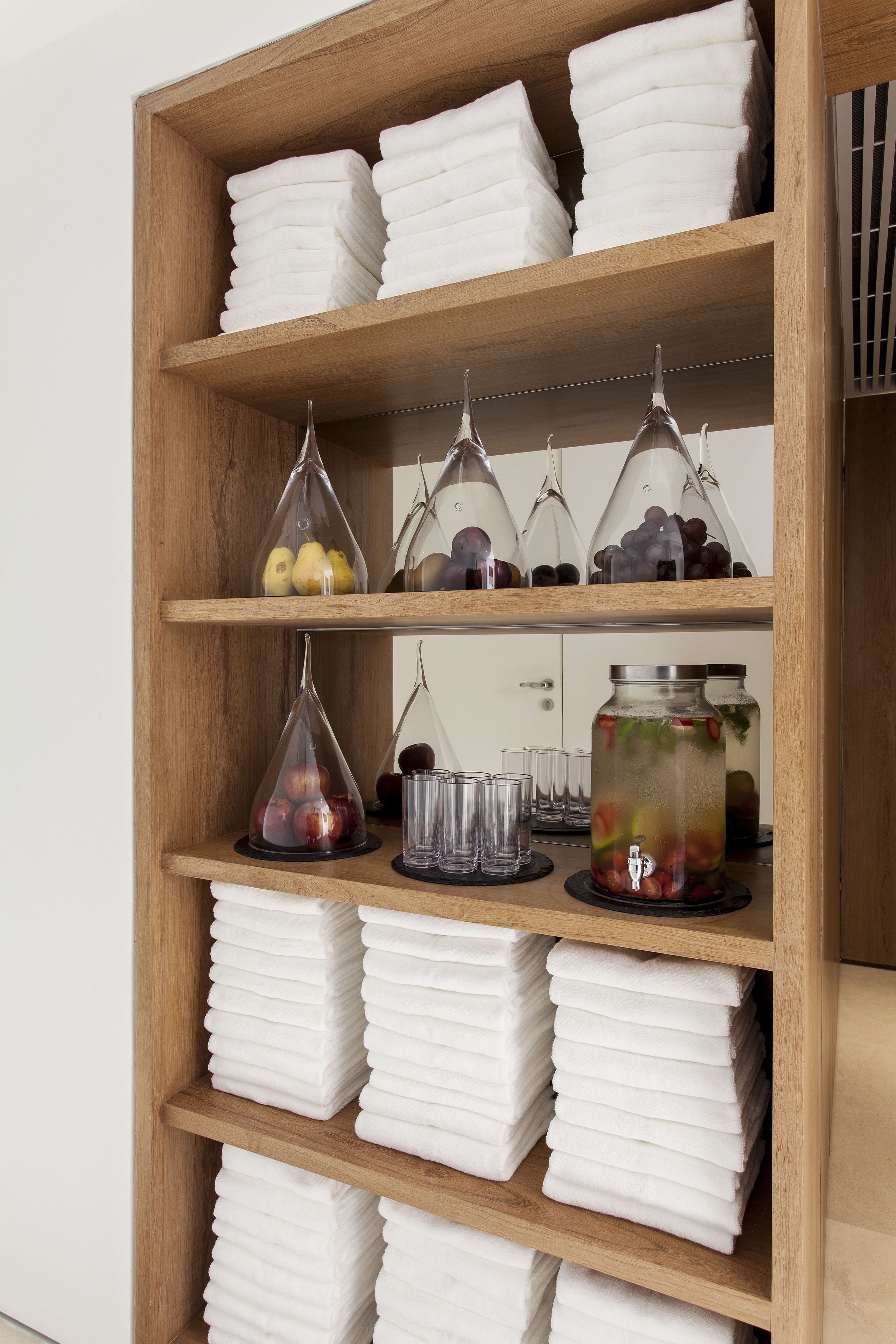 A wooden shelf with neatly stacked white towels, clear glass vases with fruits, and a glass jar with a metal lid containing a beverage.