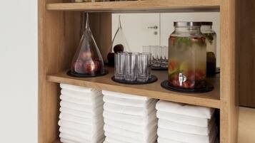 A wooden shelf with neatly stacked white towels, clear glass vases with fruits, and a glass jar with a metal lid containing a beverage.