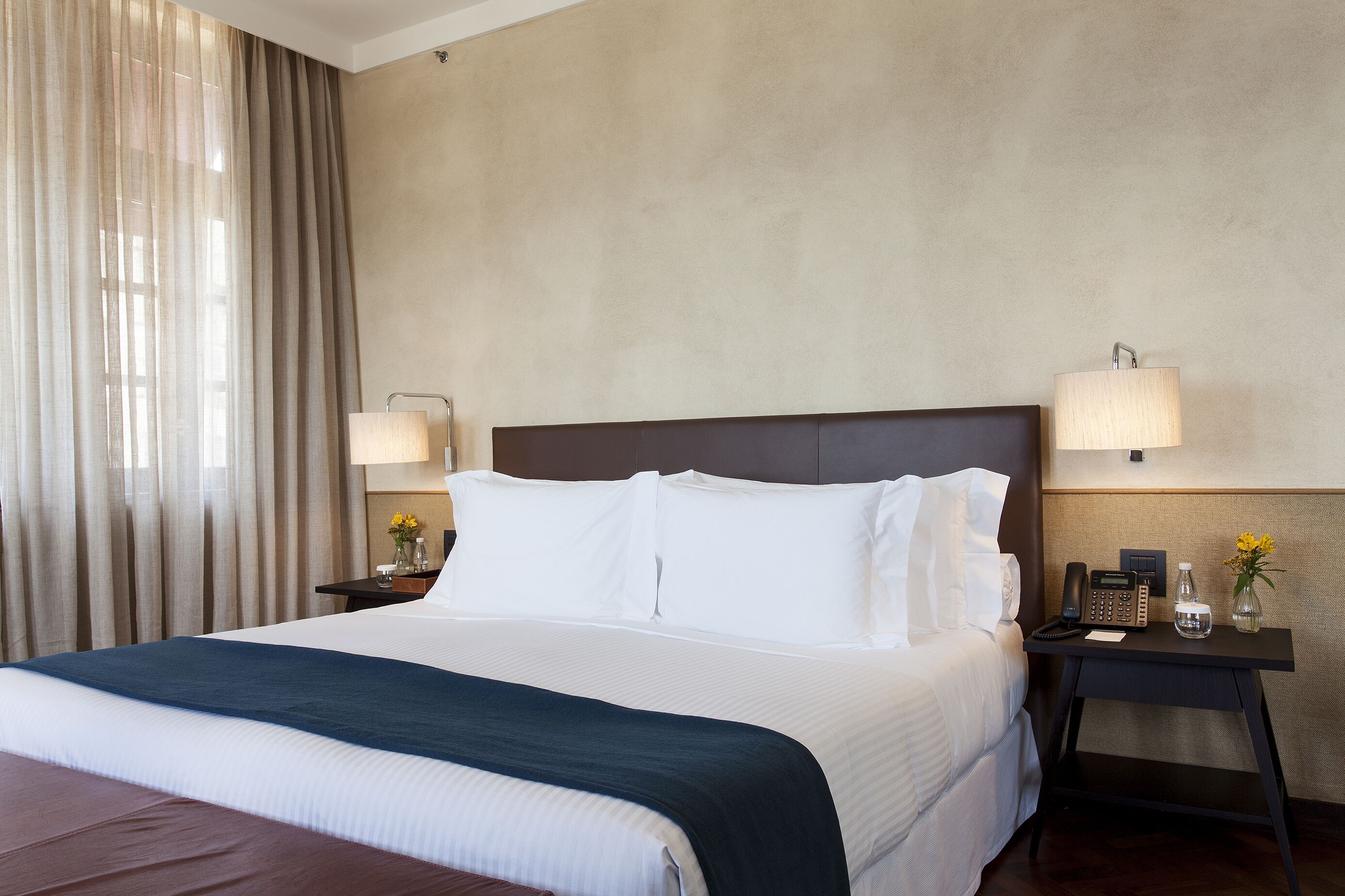 A hotel room with a large bed, two bedside tables with lamps, a telephone, and a vase with flowers.