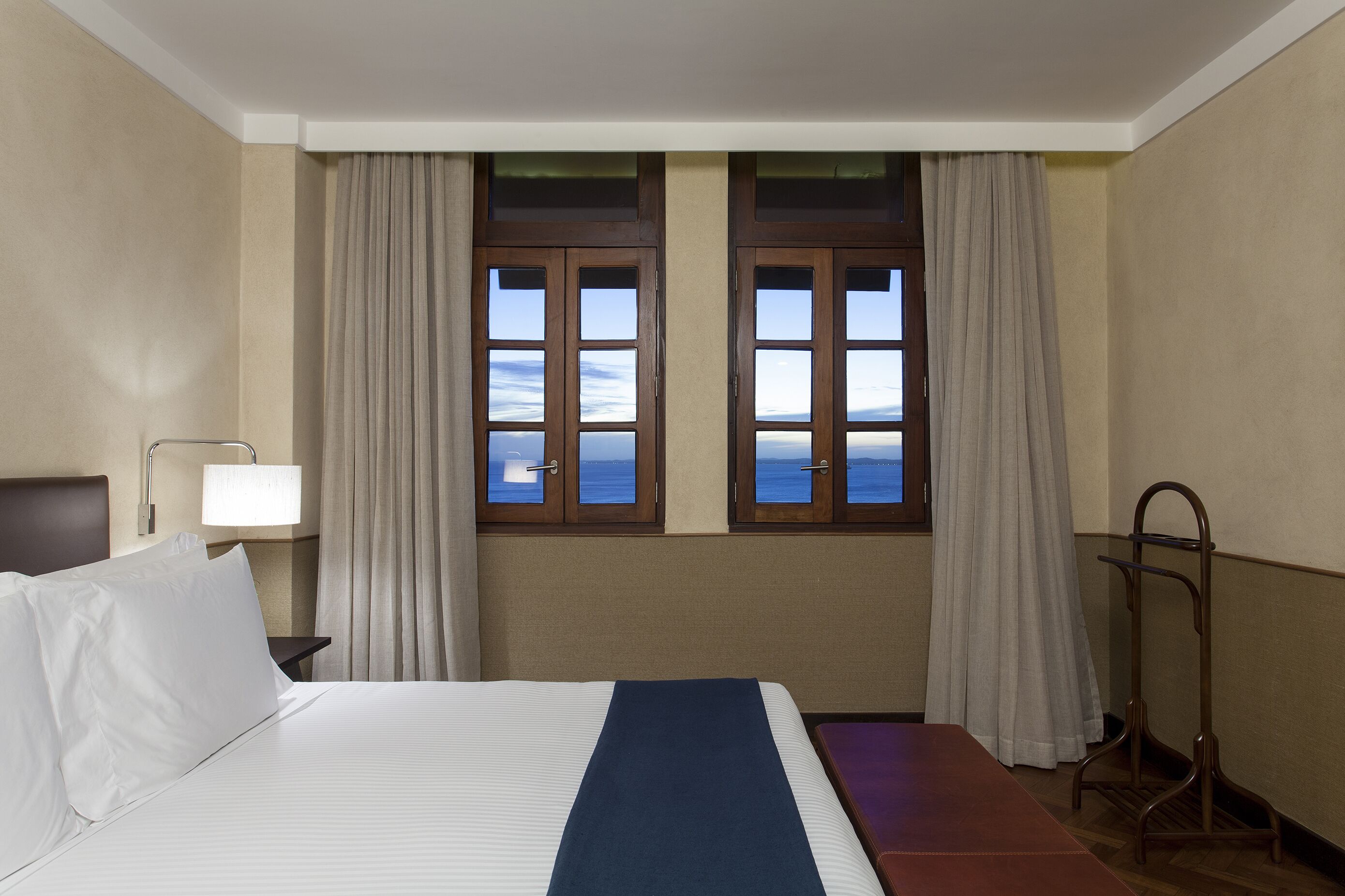 A hotel room with a bed, a nightstand, a lamp, a window with curtains, and a view of the sea.