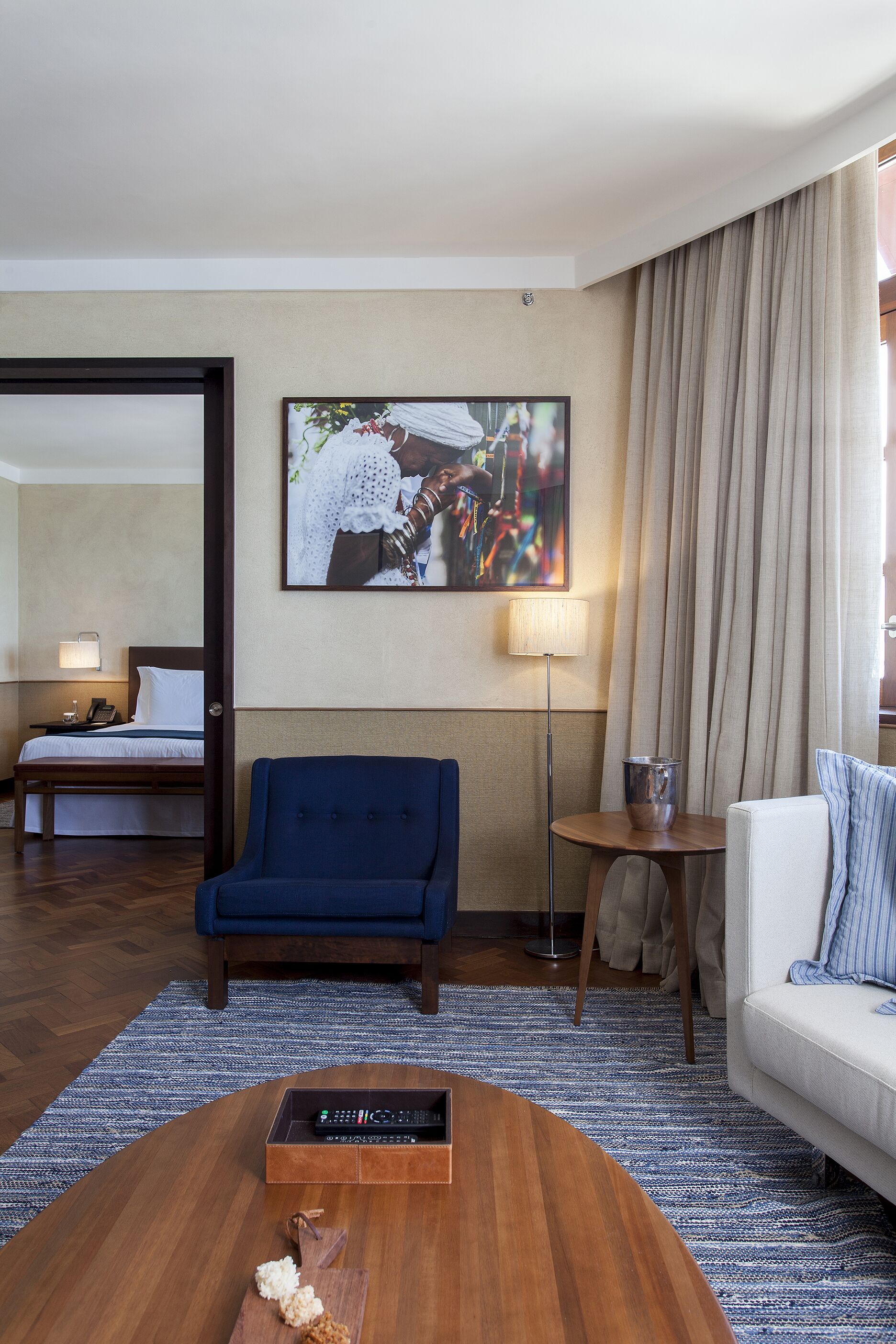 Habitació d'hotel amb un llit, un butac armchair blau, un sofà blanc, una taula de cafè, una làmpada i un quadre a la paret.
