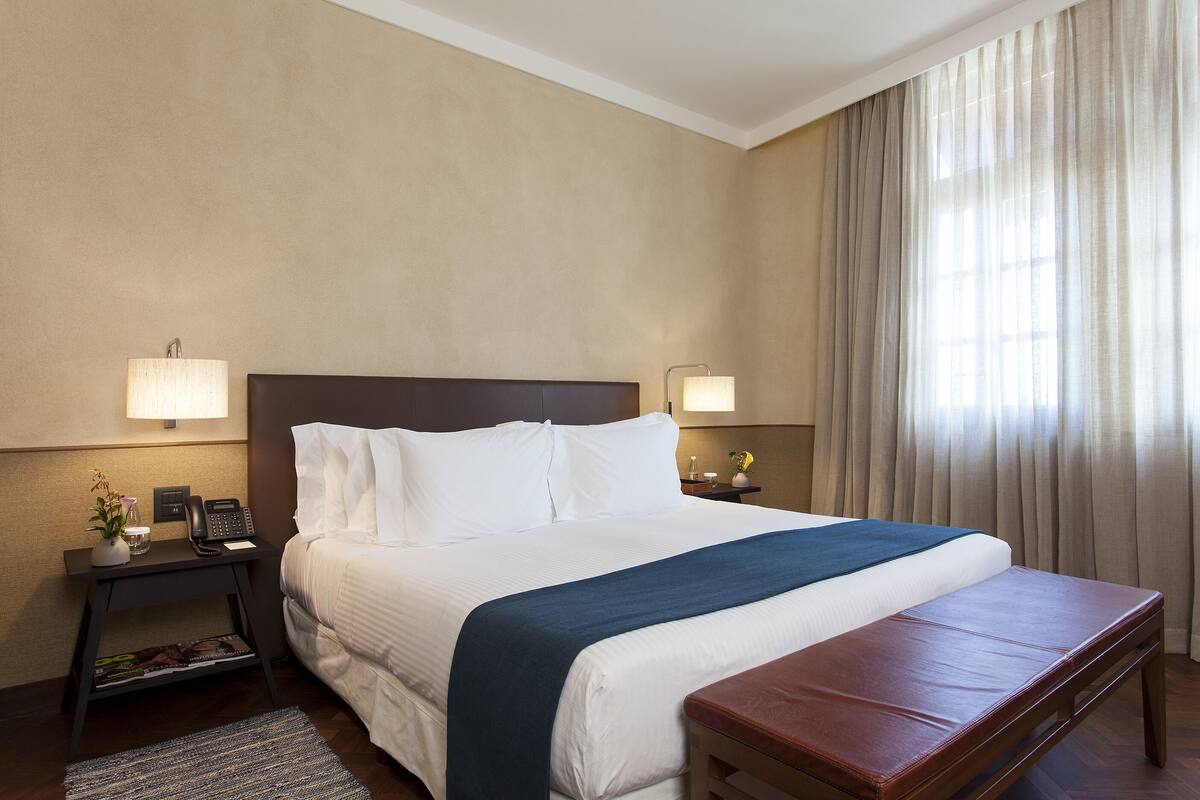 Ein Hotelzimmer mit einem großen Bett, Nachttischlampen, einem Nachttisch, einem kleinen Tisch mit einer Vase und einem Fenster mit Vorhängen.