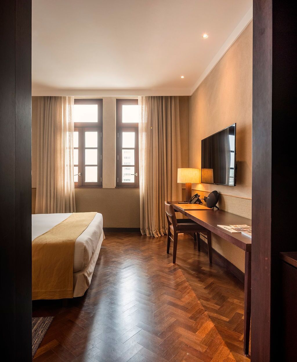 A hotel room with a bed, a desk, a chair, a lamp, a TV, and two windows with curtains.