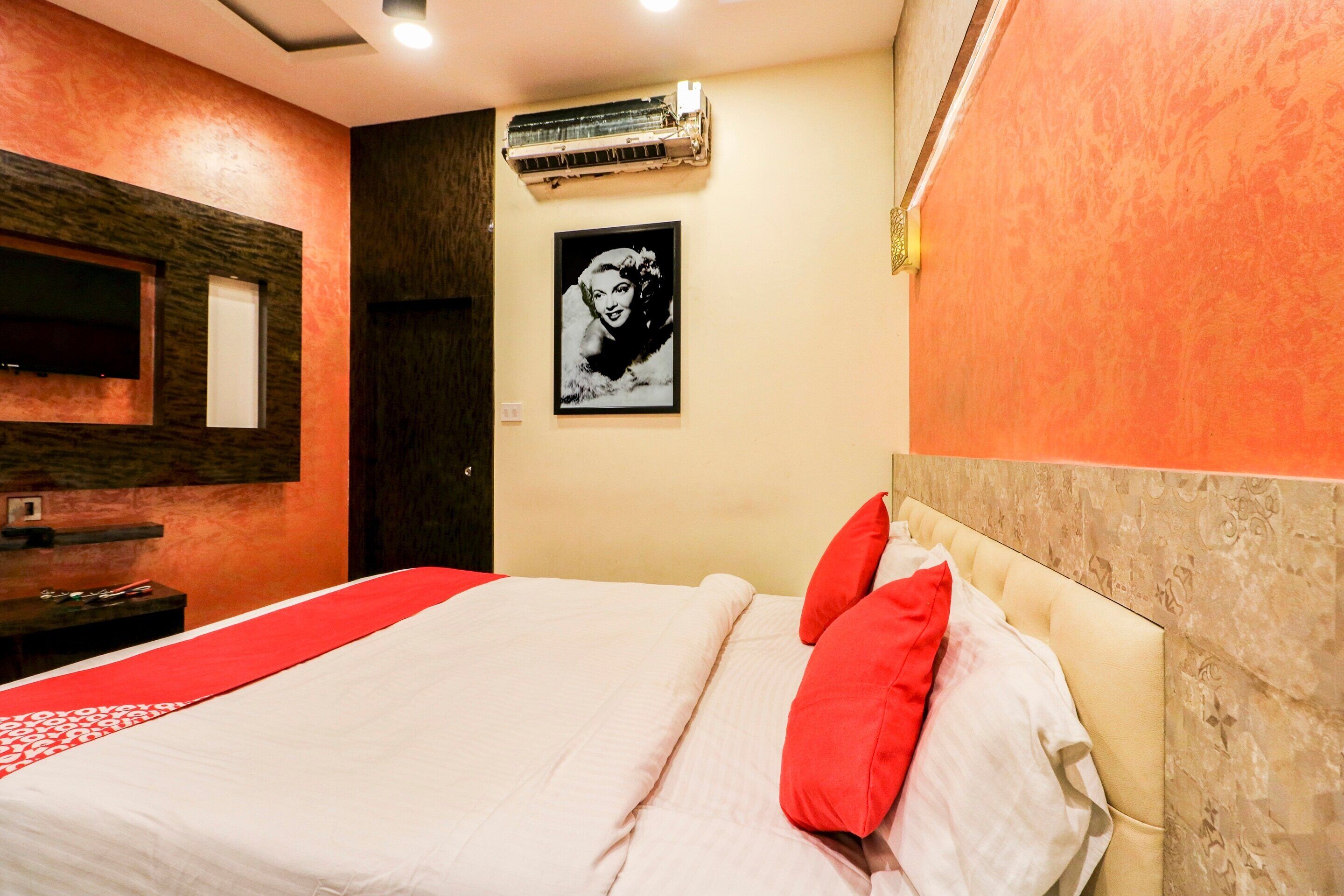 A hotel room with a bed, a red and white bedspread, a red pillow, a wall-mounted air conditioner, a framed picture, and a wooden headboard.