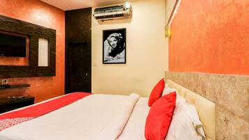A hotel room with a bed, a red and white bedspread, a red pillow, a wall-mounted air conditioner, a framed picture, and a wooden headboard.