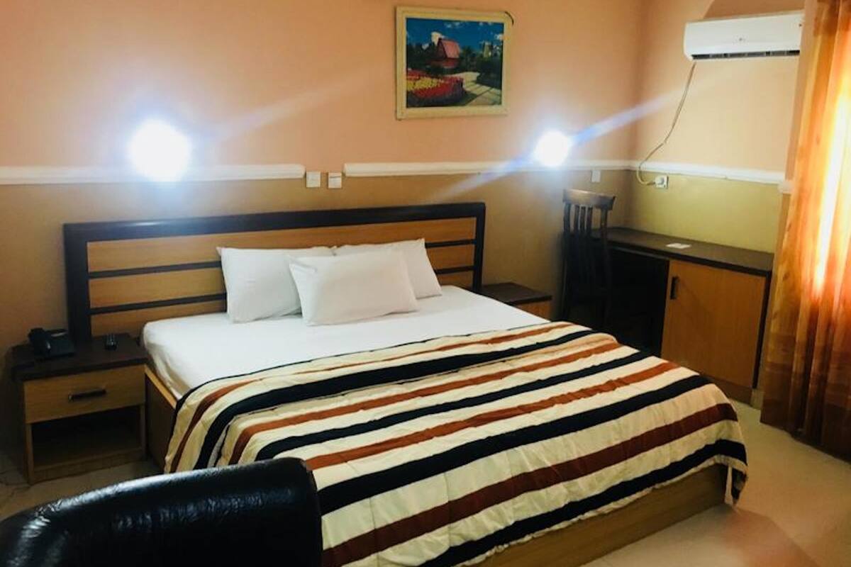 A hotel room with a bed, a nightstand, a desk, and an air conditioning unit.