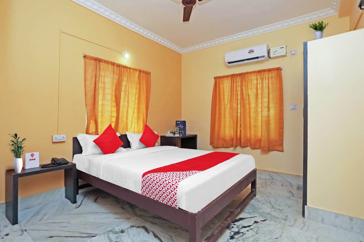 A hotel room with a bed, two nightstands, an air conditioning unit, and a potted plant.