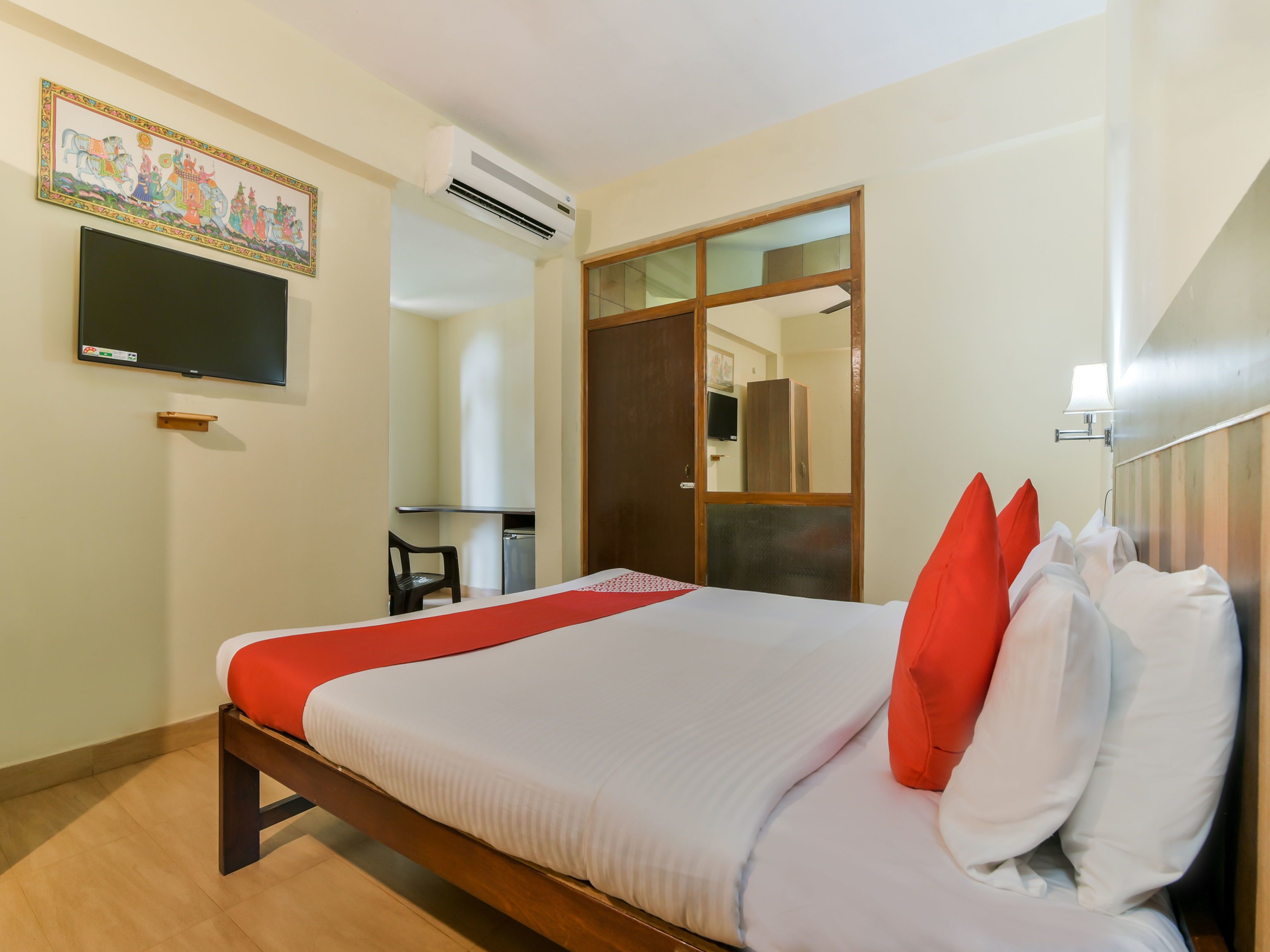 A hotel room with a bed, a TV, an air conditioning unit, a mirror, and a wooden door.