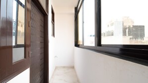 Un couloir étroit, avec une porte en bois et des fenêtres d’un seul côté, qui mène à un sol carrelé de carreaux blancs et à un mur uni.