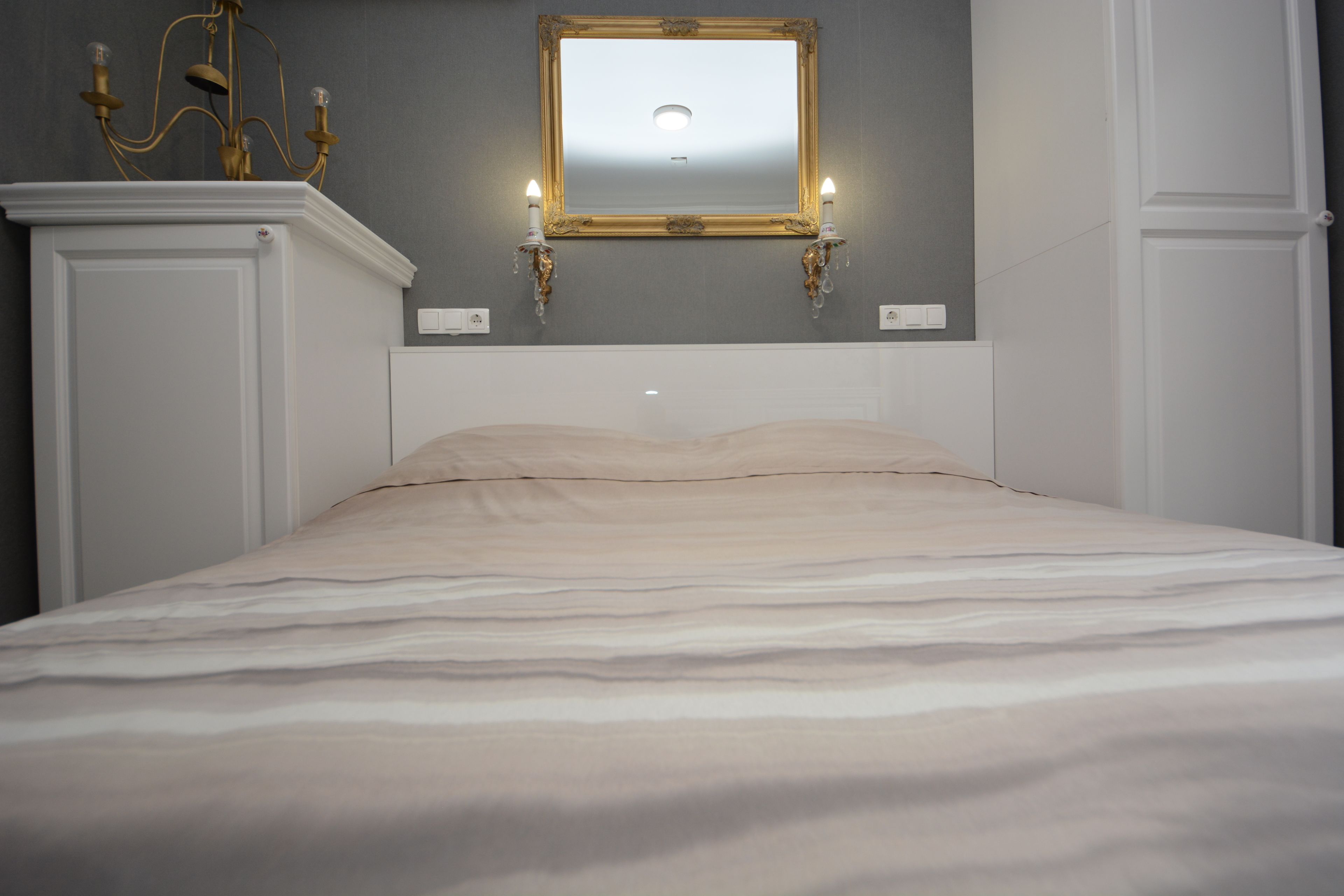 A neatly made bed with a headboard, a wall-mounted mirror with two wall sconces, and a chandelier.