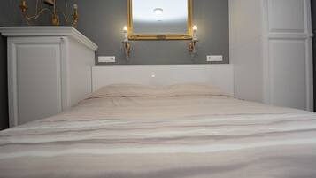 A neatly made bed with a headboard, a wall-mounted mirror with two wall sconces, and a chandelier.