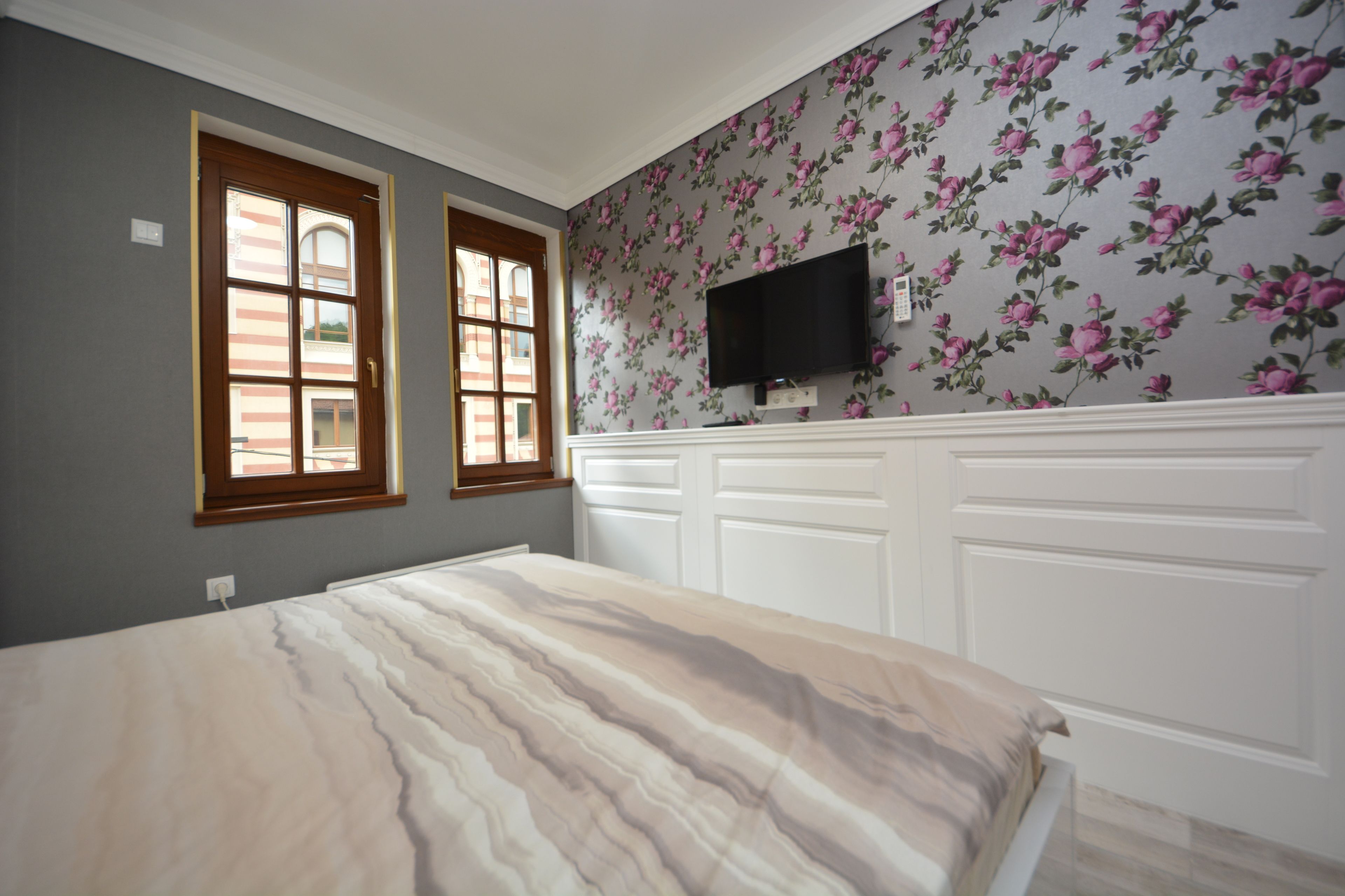Un dormitorio con cama, dos ventanas, televisor montado en la pared y un diseño de papel tapiz floral.