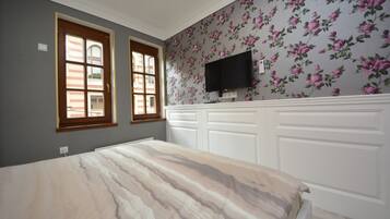 Un dormitorio con cama, dos ventanas, televisor montado en la pared y un diseño de papel tapiz floral.
