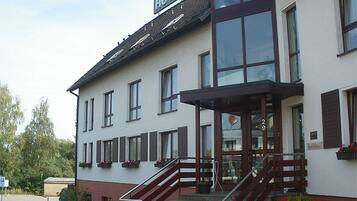 Un bâtiment à deux étages, sur lequel on peut voir une enseigne indiquant qu’il s’agit d’un hôtel. Des marches mènent à l’entrée, et il y a un petit porche avec un banc.