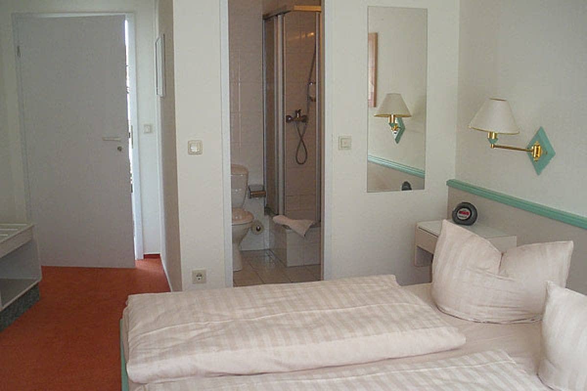 A hotel room with a bed, a bathroom with a shower, a mirror, and a nightstand with a lamp.