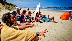 Un grupo de personas sentadas en una playa arenosa, algunas sosteniendo tablas de surf, con una tienda de campaña y un paisaje urbano al fondo.