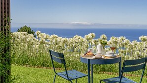 An outdoor dining area with a table set for two, surrounded by lush greenery and white flowers, overlooking a clear blue sky and distant horizon. - Quinta Inácia by Our Madeira (Calheta)