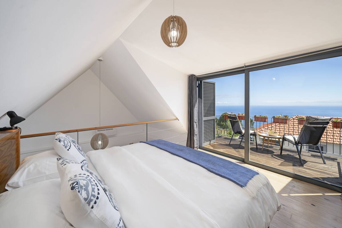 A bedroom with a large bed, a view of the sea through a sliding glass door, and a balcony with outdoor furniture.