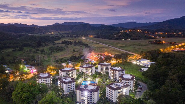 A residential area with multiple apartment buildings, a swimming pool, and a road leading to a distant hill. - Costa Linda Condo 1.3 (Jacó)