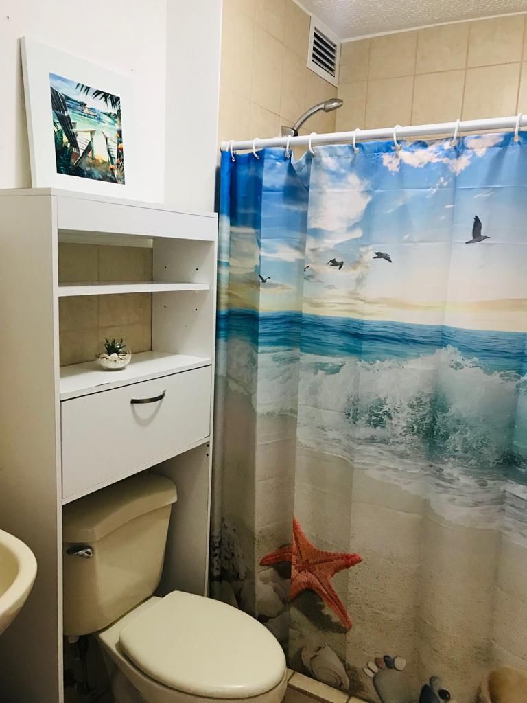 Un baño con inodoro, estante con una planta pequeña y cortina de ducha con escena de playa.