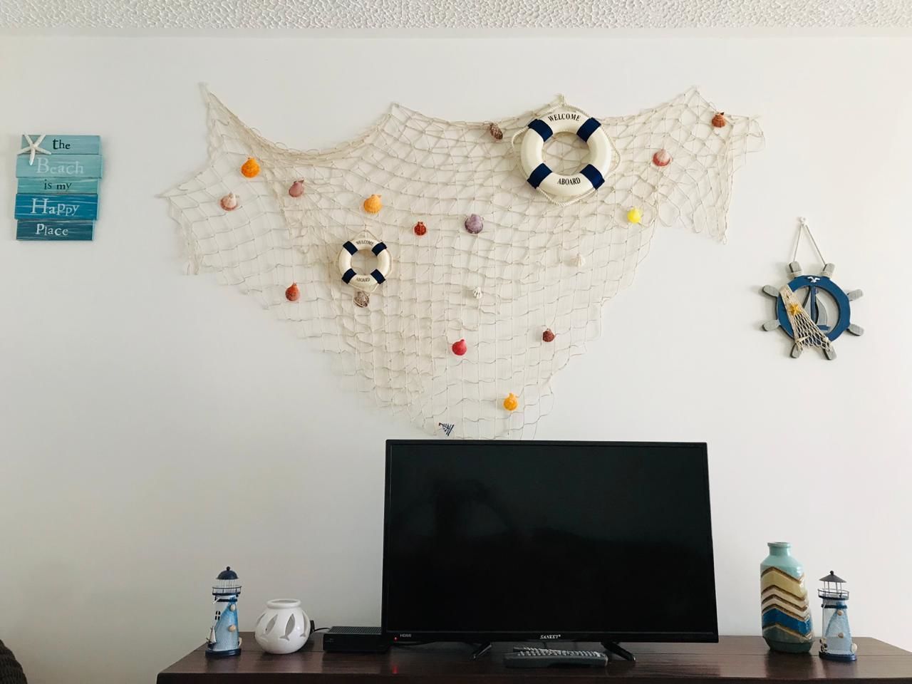 Uma televisão de tela plana sobre um suporte de madeira, um enfeite de parede com design semelhante a uma rede e vários objetos pequenos, e algumas decorações de parede incluindo uma estrela-do-mar e uma placa de boas-vindas.