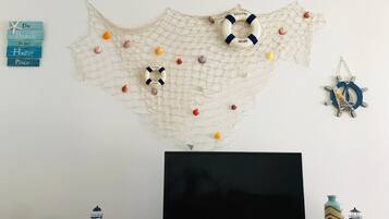 Uma televisão de tela plana sobre um suporte de madeira, um enfeite de parede com design semelhante a uma rede e vários objetos pequenos, e algumas decorações de parede incluindo uma estrela-do-mar e uma placa de boas-vindas.