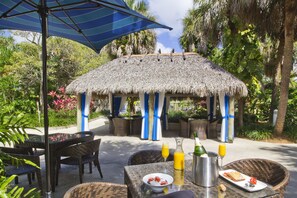 A thatched-roof outdoor dining area with wicker furniture, a table with a bottle of champagne, a pitcher, and a glass of orange juice. - Miami Lakes Hotel and Golf (Miami Lakes)
