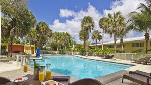 Un espace aménagé au bord de la piscine, comprenant un coin repas avec une table et des chaises, une bouteille de champagne et un verre de jus d’orange.