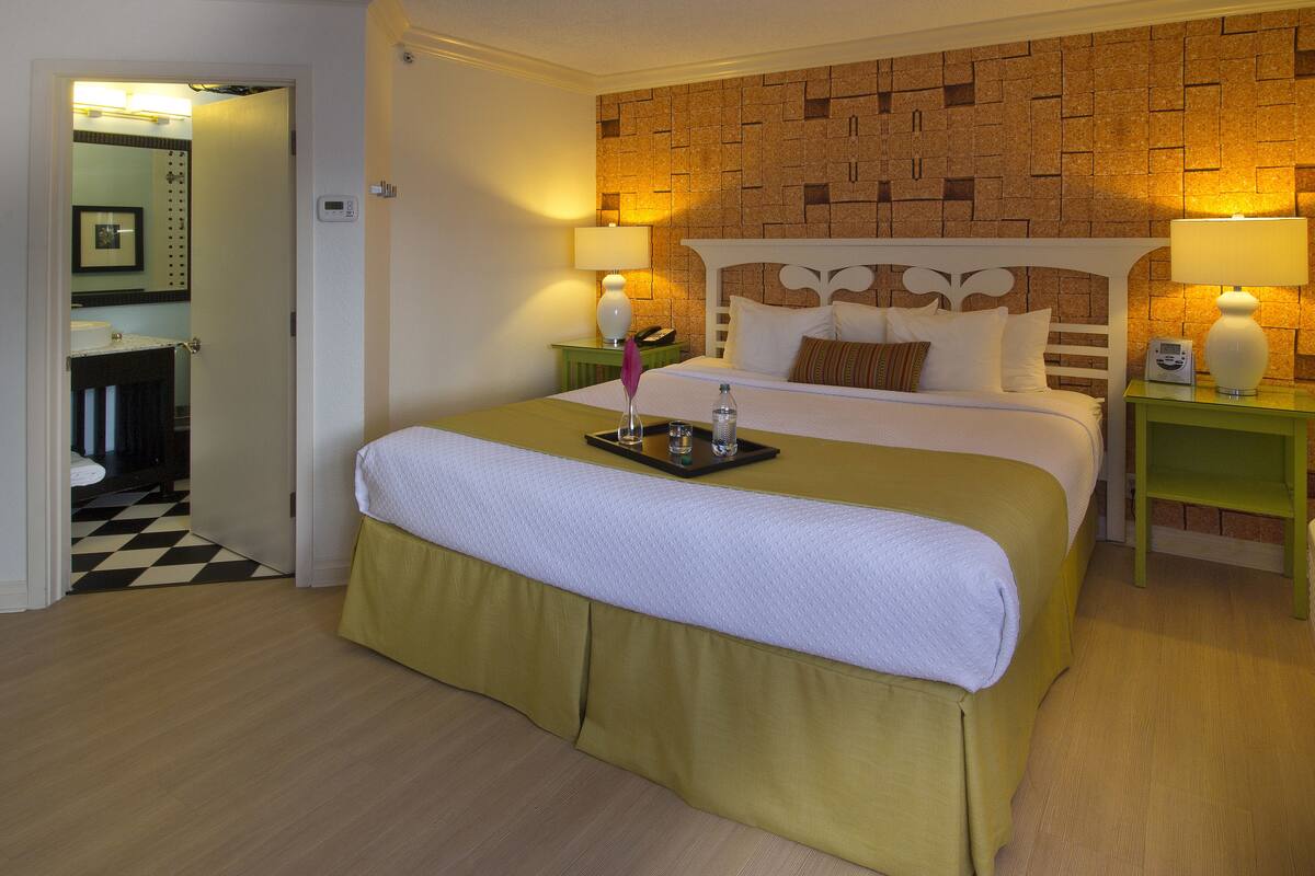 A hotel room with a large bed, two bedside tables with lamps, a green nightstand, a bathroom visible through an open door, and a checkered floor.