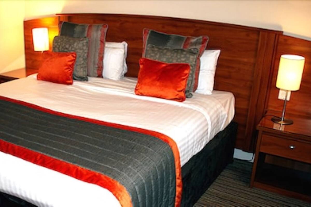 A hotel room with a large bed, multiple pillows, a wooden headboard, bedside lamps, and a nightstand.