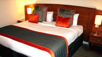 A hotel room with a large bed, multiple pillows, a wooden headboard, bedside lamps, and a nightstand.
