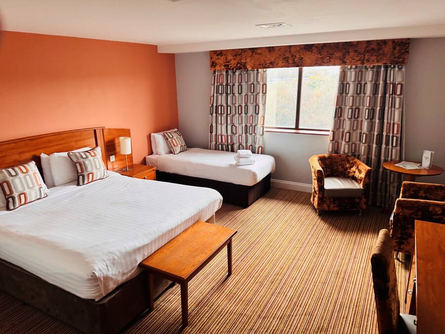Een hotelkamer met twee bedden, een houten hoofdeinde, een bankje, een stoel, een tafeltje en een raam met geruit gordijn.