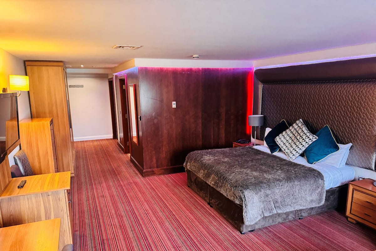 A hotel room with a large bed, a wooden desk, a wardrobe, and a red carpeted floor.