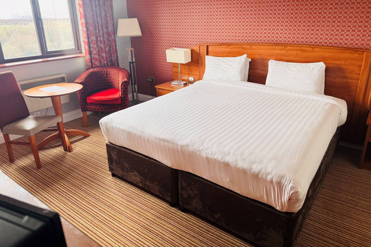 A hotel room with a large bed, a small round table, a red chair, a lamp, and a window with curtains.