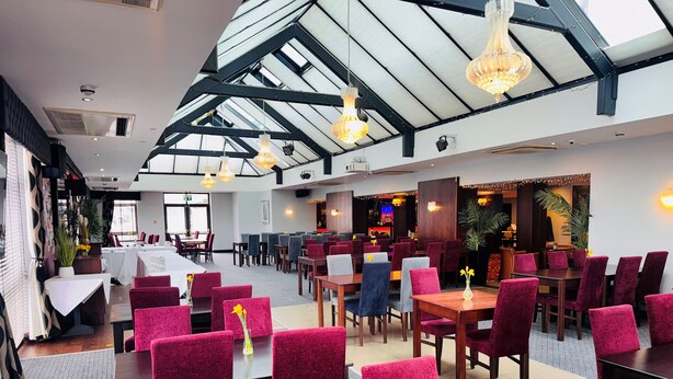 A spacious restaurant with high ceilings and natural light. Tables are arranged in clusters, some with red upholstered chairs.
