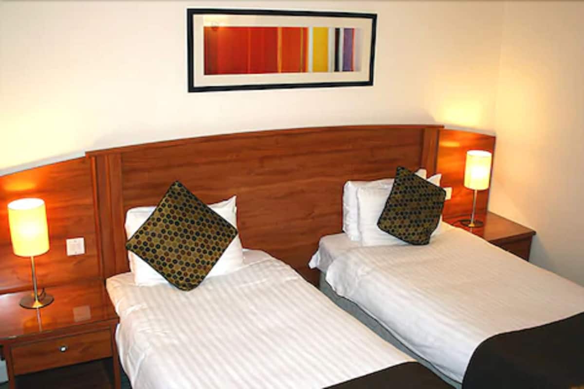 A hotel room with two beds, each with a wooden headboard, white linens, and patterned pillows. There are bedside lamps and a framed abstract painting on the wall.