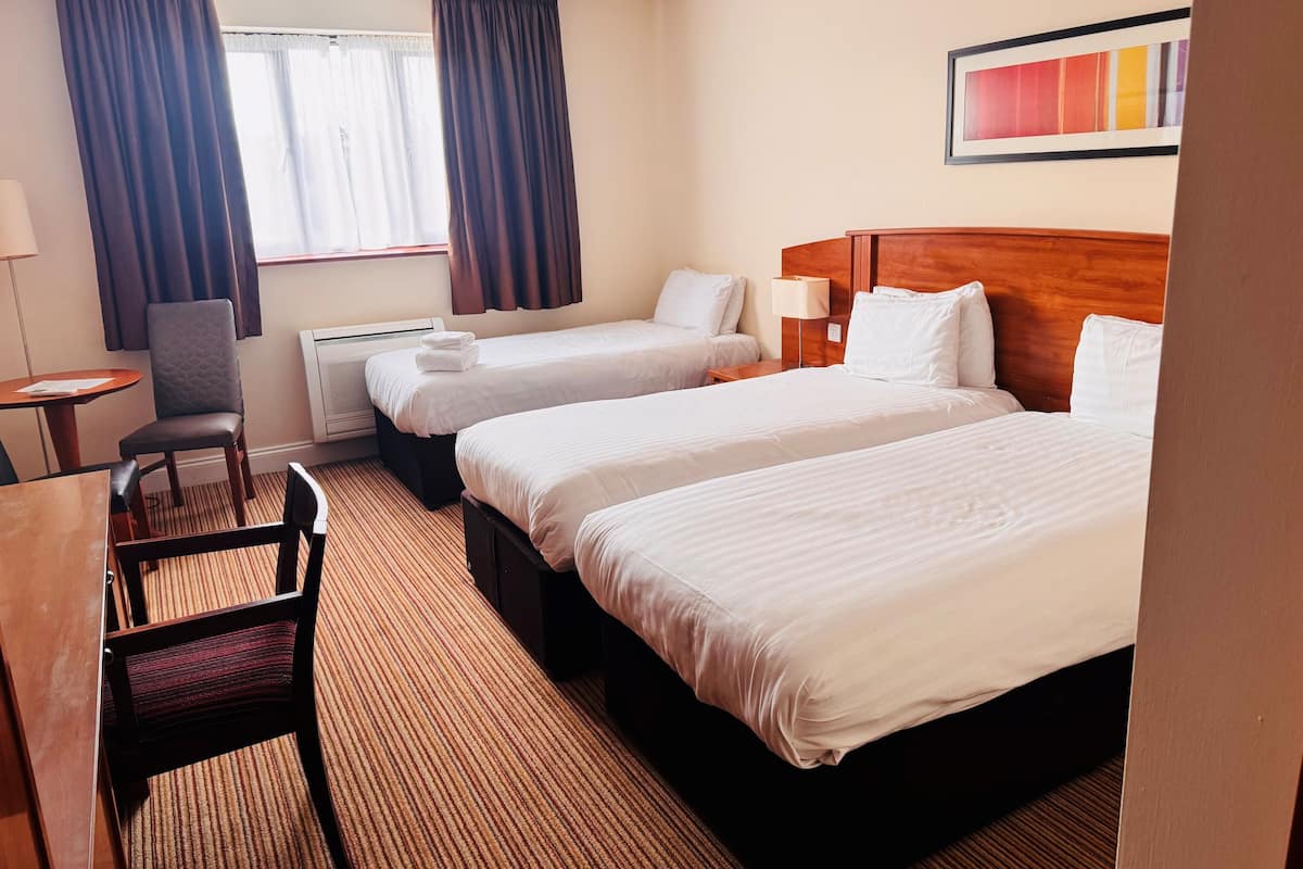 A hotel room with two single beds, a desk with a chair, a small table, and a window with curtains.
