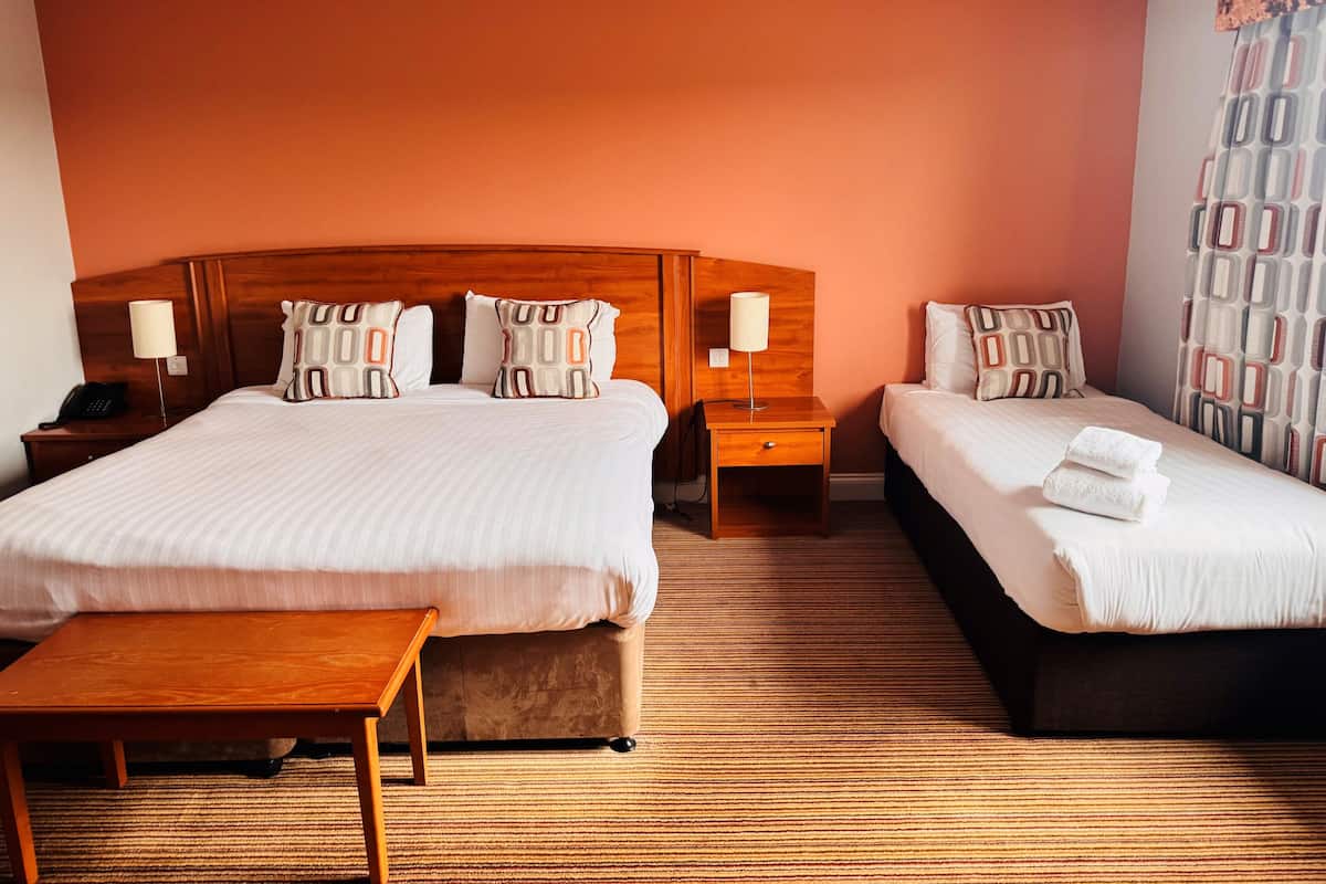 A hotel room with two beds, a wooden headboard, a bedside table with a lamp, and a small wooden table.