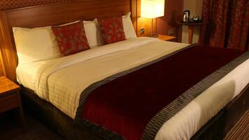 A hotel room with a large bed, a wooden headboard, a bedside table with a lamp, and a small table with a kettle and cups.