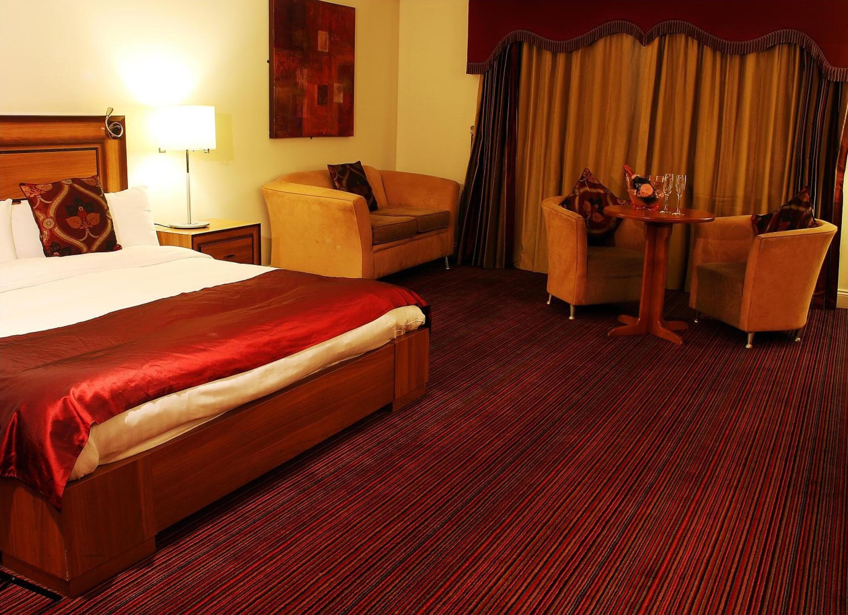 Habitación de hotel con una cama grande, un sofá, una mesita con sillas y vistas al exterior a través de cortinas.