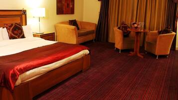 Habitación de hotel con una cama grande, un sofá, una mesita con sillas y vistas al exterior a través de cortinas.