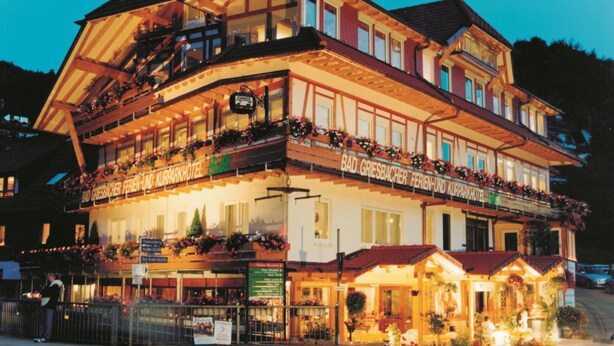 Ein mehrstöckiges Hotel im traditionellen alpinen Baustil, mit Balkonen, die mit Blumenkästen geschmückt sind, und einem Restaurant im Erdgeschoss.